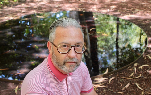 A white man with cropped grey hair and brown rimmed glasses sits in natural sunlight, in front of a glass table in an outdoor environment. He wears a pink shirt.