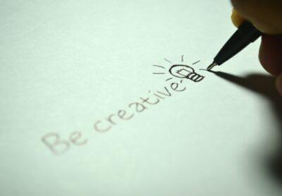 a hand writes the words 'be creative' onto a piece of white paper, with a lightbulb doodle to punctuate the sentence