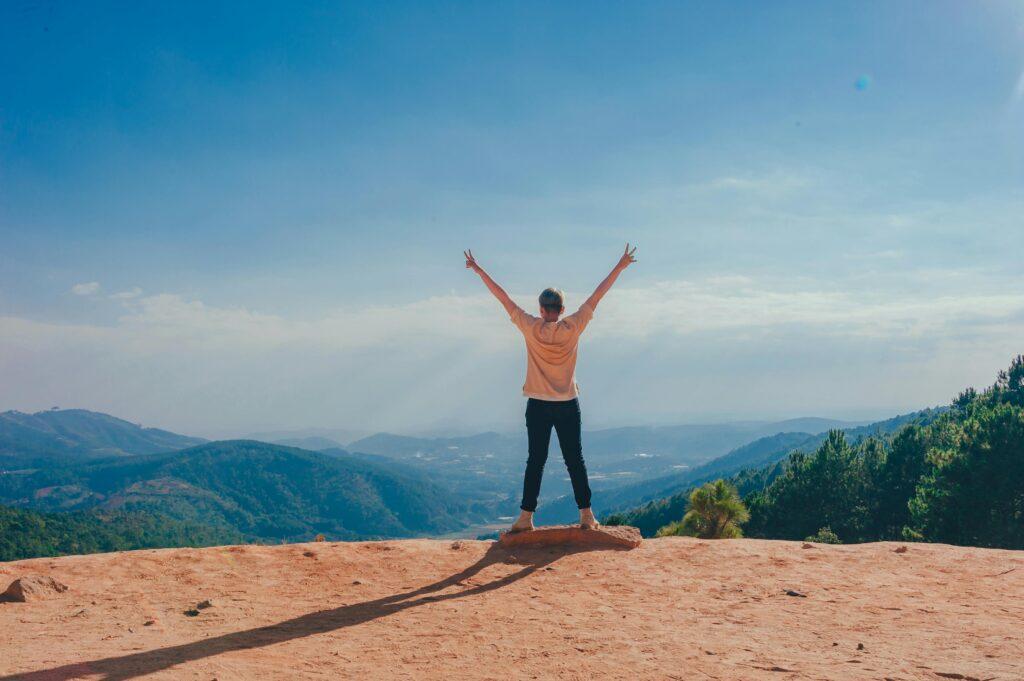 A man stands triumphantly on the side of a mountain looking out onto a gorgeous sunlit view